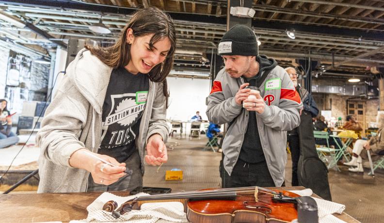 A woman with a staff shirt is explaining how to repair and clean a violin. A man holding a coffee cup is watching her work.