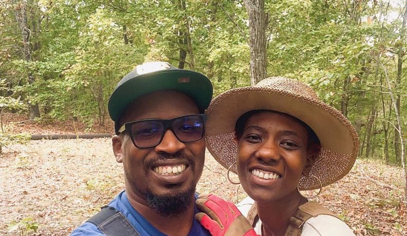 Osei Doyle and Brendalyn King are dressed in overalls and t-shirts, wearing brimmed hats. They are taking a selfie outside, with Brendalyn holding onto Osei's shoulder.