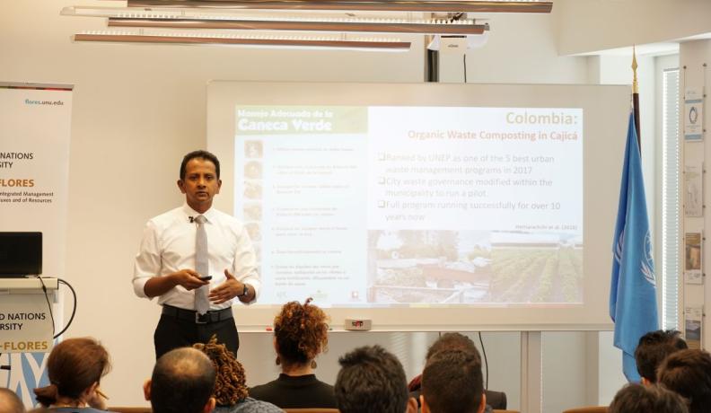 Hiroshan Hettiarachchi is giving a seminar, speaking to a class of diverse people that are all facing him. Behind Hettiarachchi is a powerpoint and to the far right is the UN flag.