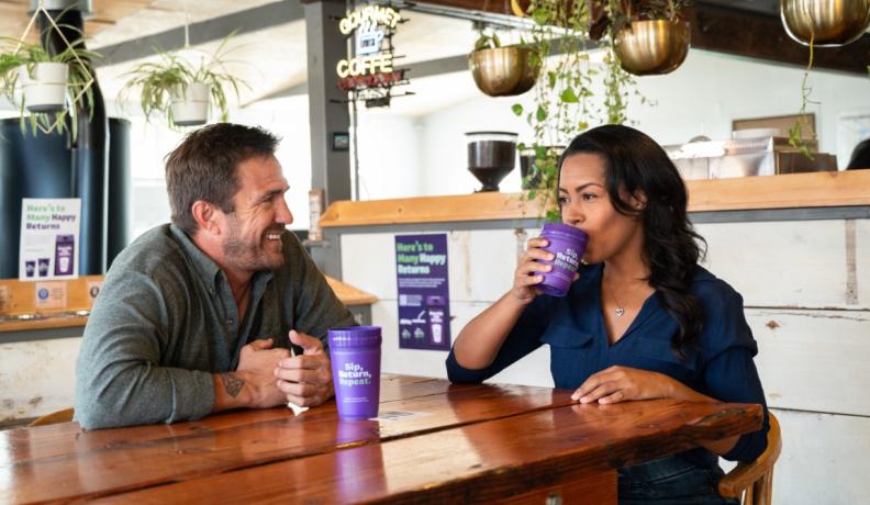Two people, a white man and a mixed descent woman, sit at a coffee table in a cafe. He is looking at her and smiling. She is taking a sip from a reusable purple cup.