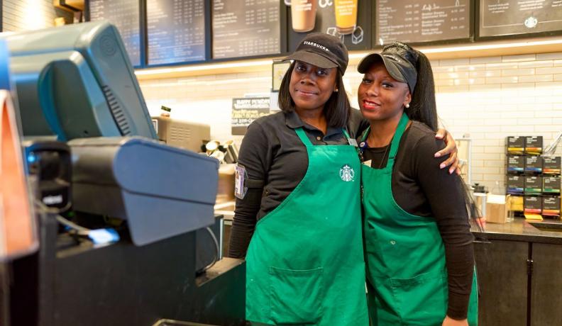 two Black women Starbucks baristas are smiling at the camera. One has her arm wrapped another the other in a side hug. They look prepared and ready for work.