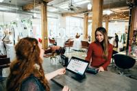 red-haired salon worker with a big smile standing behind counter accepting payment from a woman customer in a red sweater with a big smile, businesses that do a sustainability audit are on their way