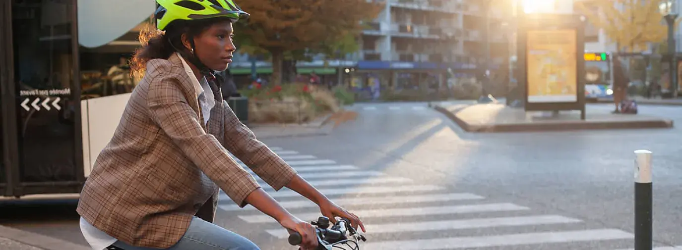 Black woman in a brown blazer, flared jeans, and a neon green helmet riding a gray bike through city streets.