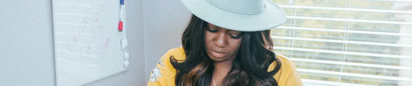 A Black woman sits at a desk in front of a laptop. She's wearing a wide-brimmed white hate and yellow floral blouse, she's looking down at the laptop. On the desk are various items - a picture frame, a box, a statue, a notebook. On the wall next to her is a whiteboard with scribbles on it and behind her is a window. Black-Owned Businesses for Black History Month. Juneteenth.