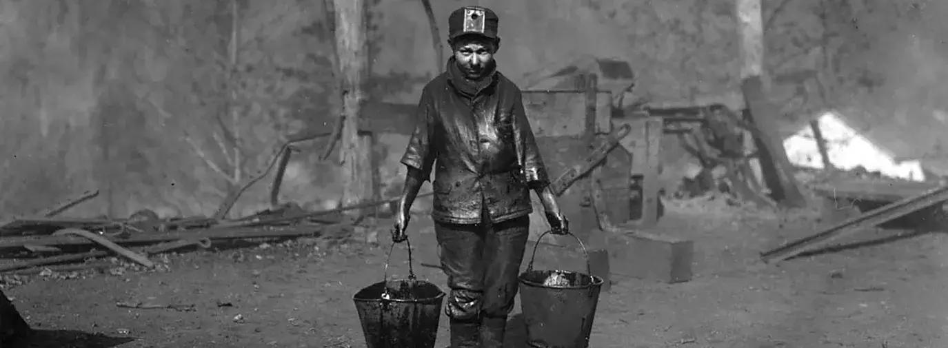 A black and white photo of a child under the age of 12 carrying two grease pails. He is covered in grease on his body. He stands alone in a bare and gray construction site on a hill.