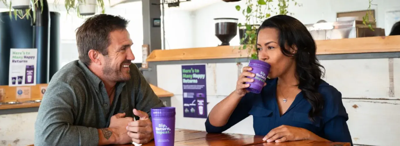 Two people, a white man and a mixed descent woman, sit at a coffee table in a cafe. He is looking at her and smiling. She is taking a sip from a reusable purple cup.
