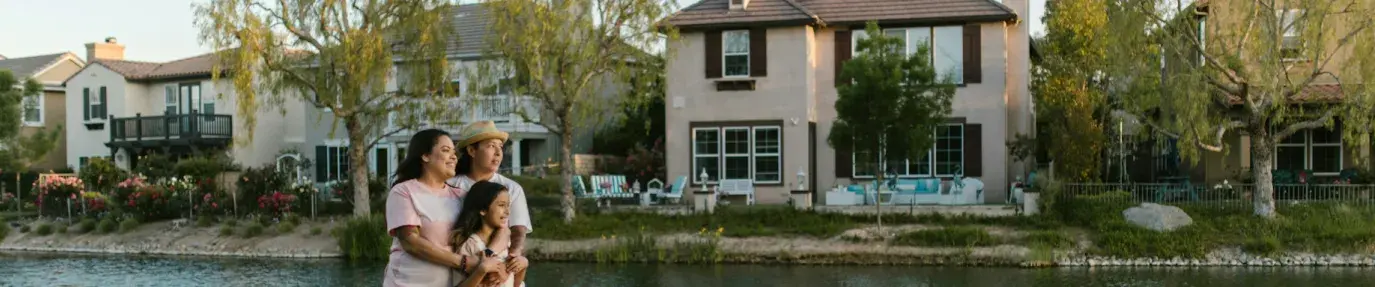 Two women stand with their arms around a young girl, their daughter, at the edge of a small lake with homes and trees in the background. Clean energy for all.