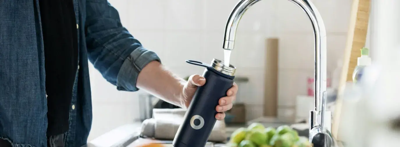 white person wearing a denim shirt and refilling a black metal water bottle in a kitchen sink.