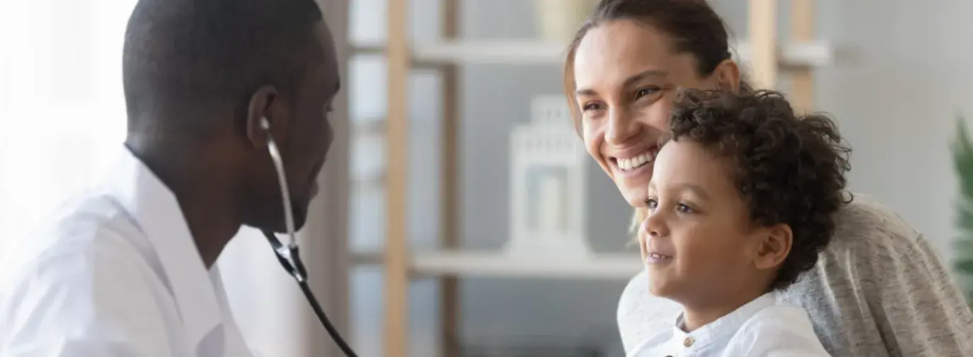 A young black doctor holds a stethoscope up to a mixed child's heart. The mom has the child on her lap. Both are smiling at the doctor.